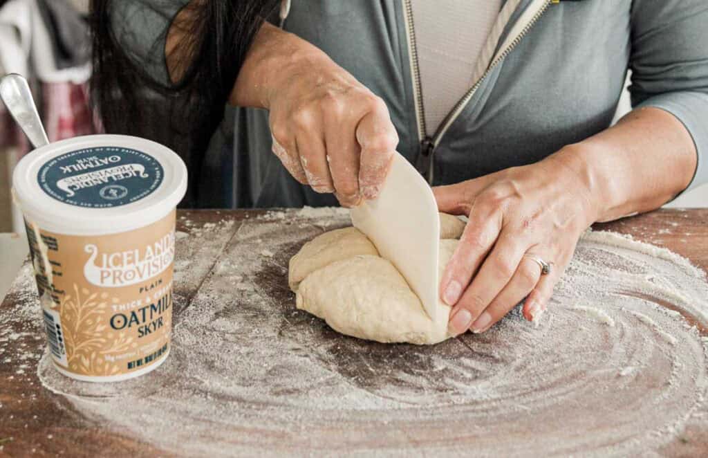 A person kneads dough on a floured surface using a white scraper. A container of Icelandic Provisions plain oatmeal skyr is nearby on the wooden table. The person is wearing a gray hoodie with a white shirt underneath.